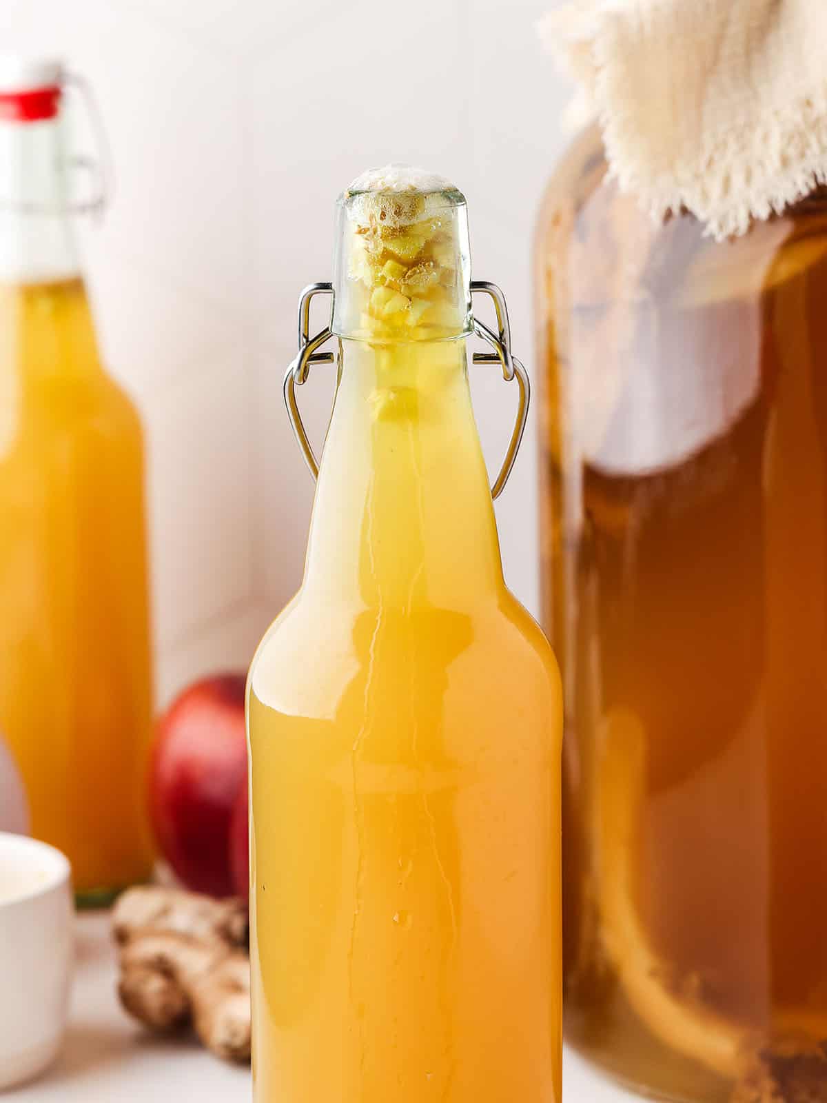 Bottles of apple ginger kombucha next to a gallon jar of regular kombucha with cheesecloth on it. 