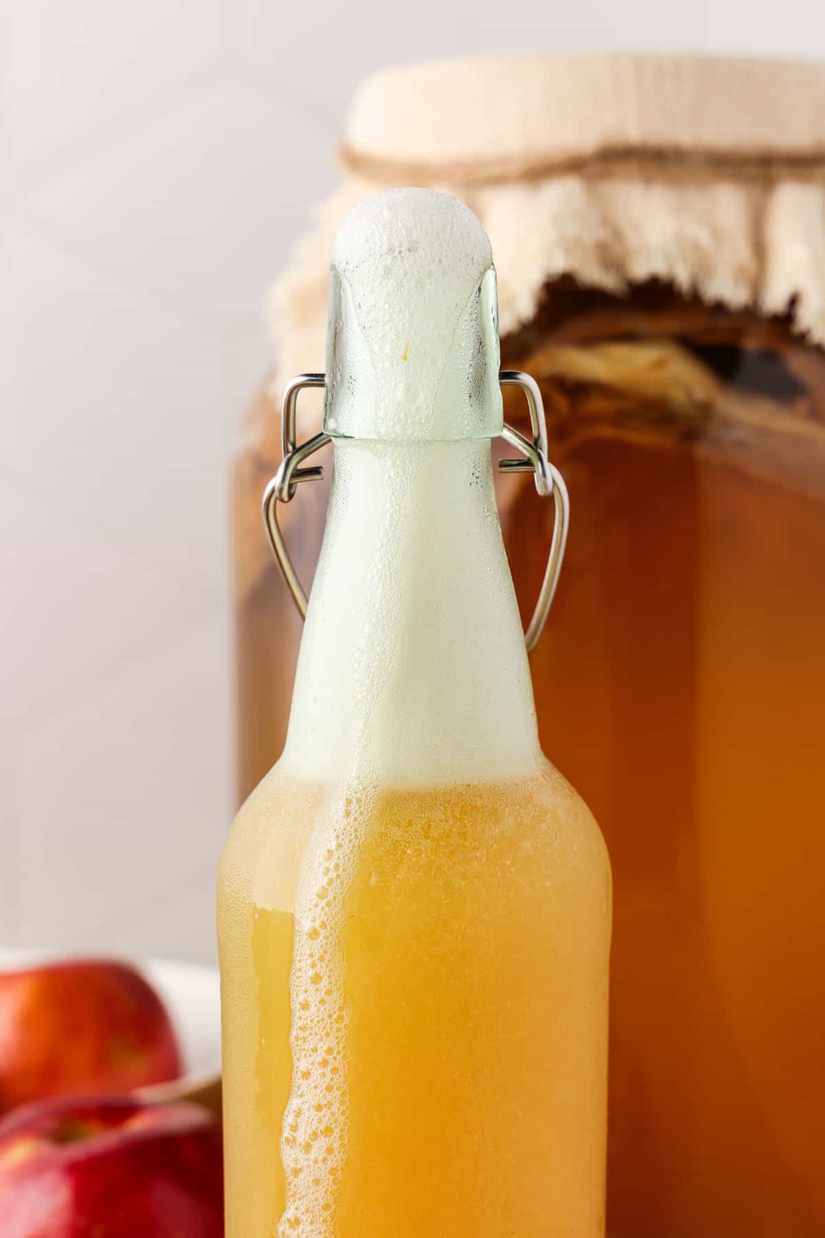 An open bottle of apple ginger kombucha with fizz up to the top of the bottle. With a big jar of regular kombucha and fresh apples in the background. 