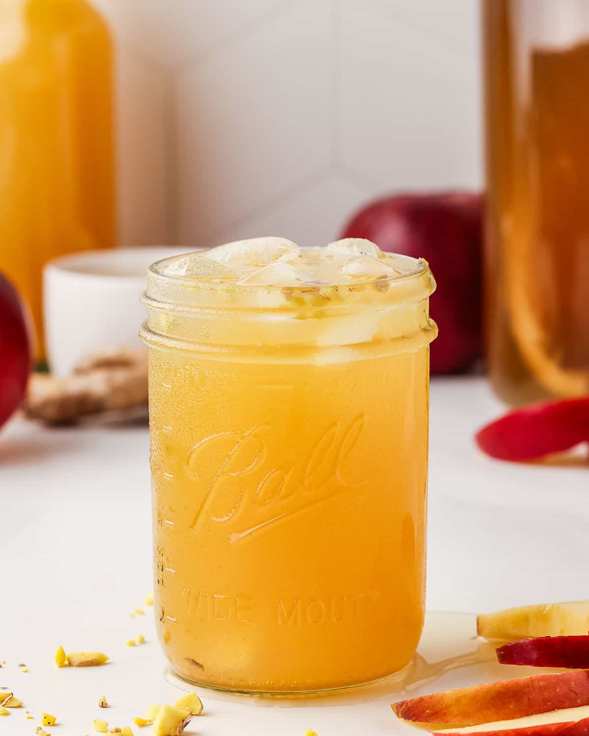 A glass of apple ginger kombucha with ice, surrounded by minced ginger and sliced apples. 