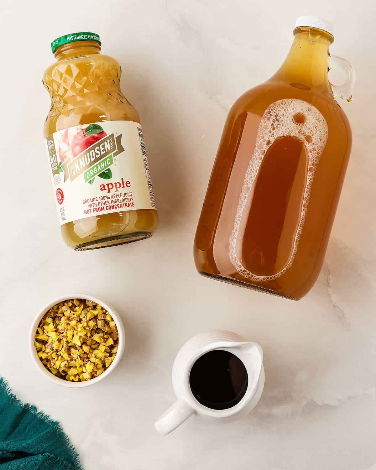 Ingredients for making apple ginger kombucha on a countertop, a bottle of kombucha, a bottle of apple juice, a bowl of minced ginger, and a dish of maple syrup. Top view. 