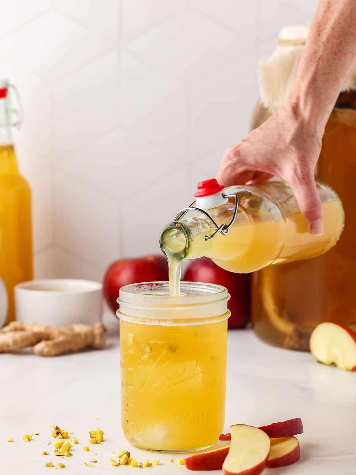 Apple ginger kombucha pouring into a glass surrounded by fresh sliced apples and ginger root. 