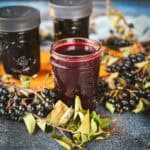 A dark purple oxymel in a jar with aronia berries and leaves surrounding.