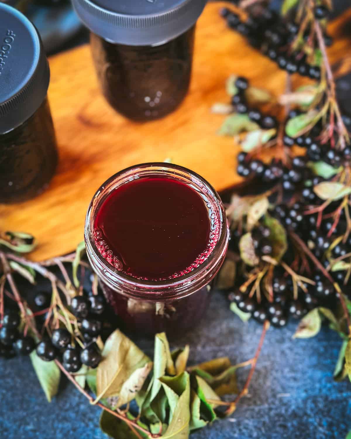 A jar of aronia oxymel with 2 more in the background, surrounded by fresh aronia plants, top view. 