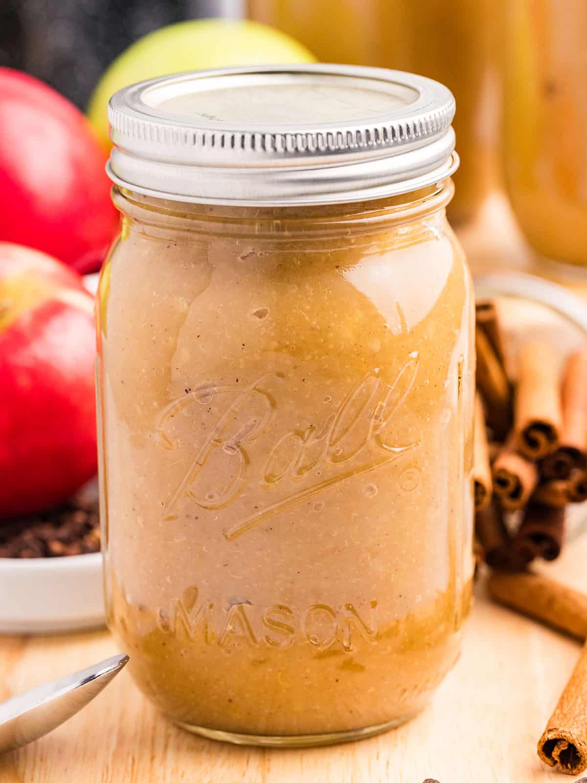 A jar of canned spiced applesauce on a wood surface, surrounded by cinnamon sticks and fresh whole apples. 