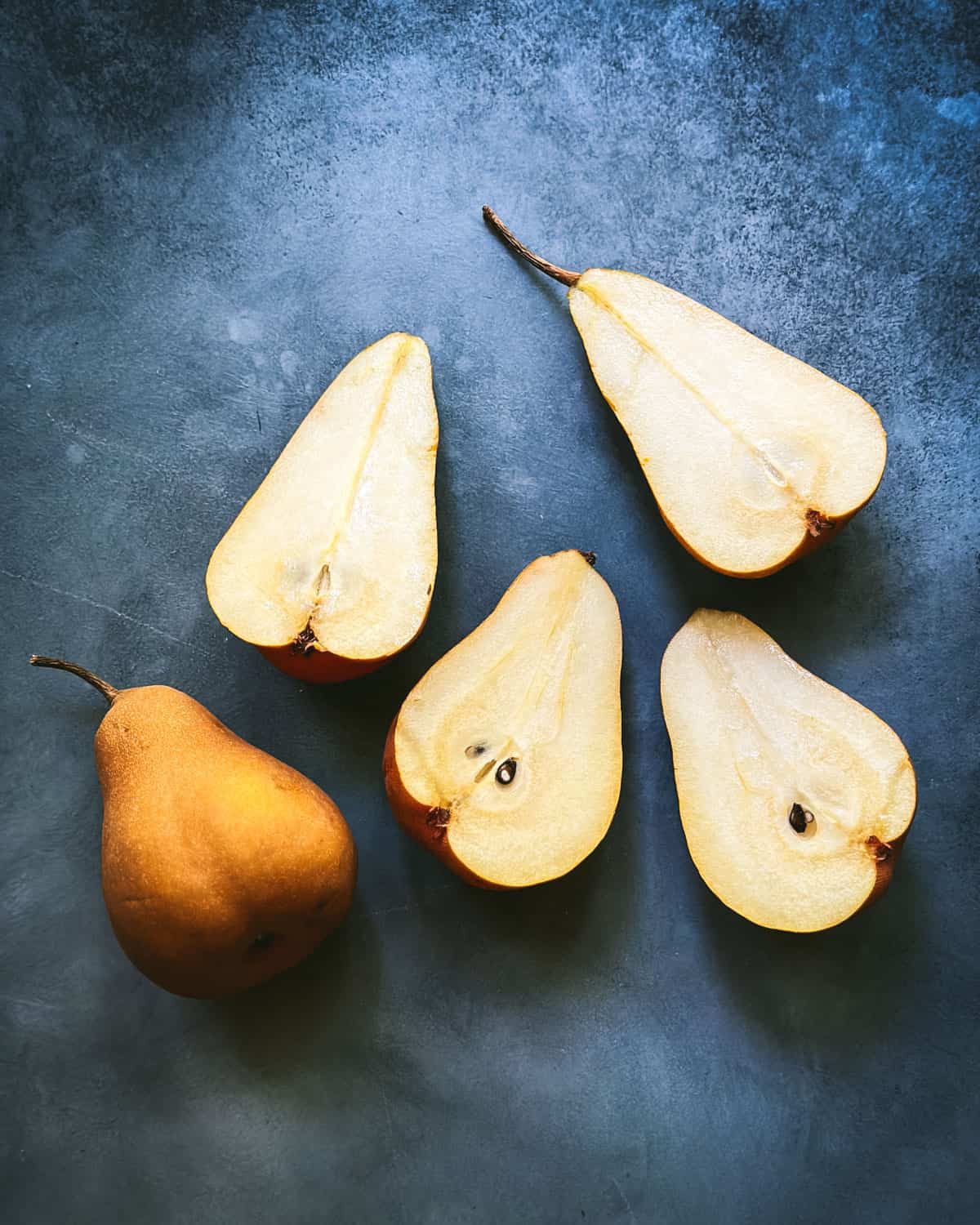 Halved pears and one whole one on a moody blue surface with natural lighting. Top view. 