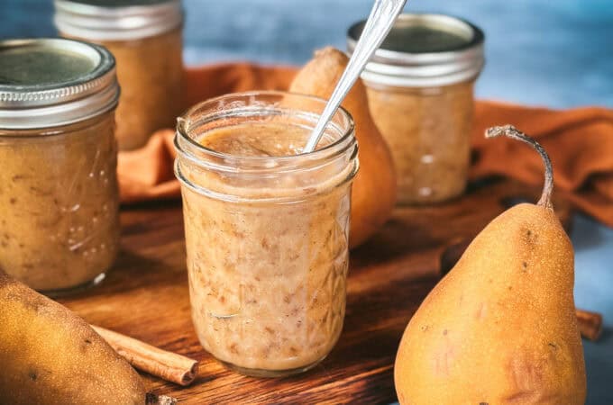 An open jar of pear butter with a spoon in it, on a wood cutting board surrounded by fresh whole pears, cinnamon sticks, and closed jars of pear butter.