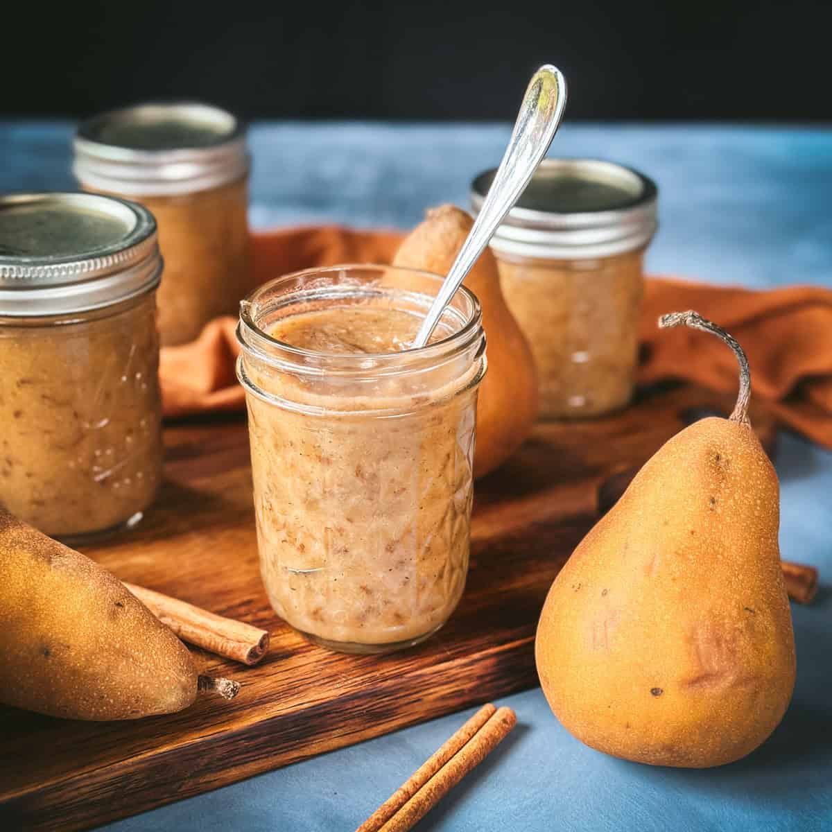 An open jar of pear butter with a spoon in it, on a wood cutting board surrounded by fresh whole pears, cinnamon sticks, and closed jars of pear butter.