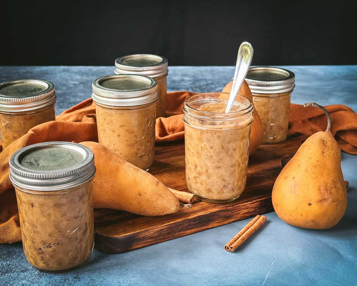 An open jar of pear butter with a spoon in it, on a wood cutting board surrounded by fresh whole pears, cinnamon sticks, and closed jars of pear butter.