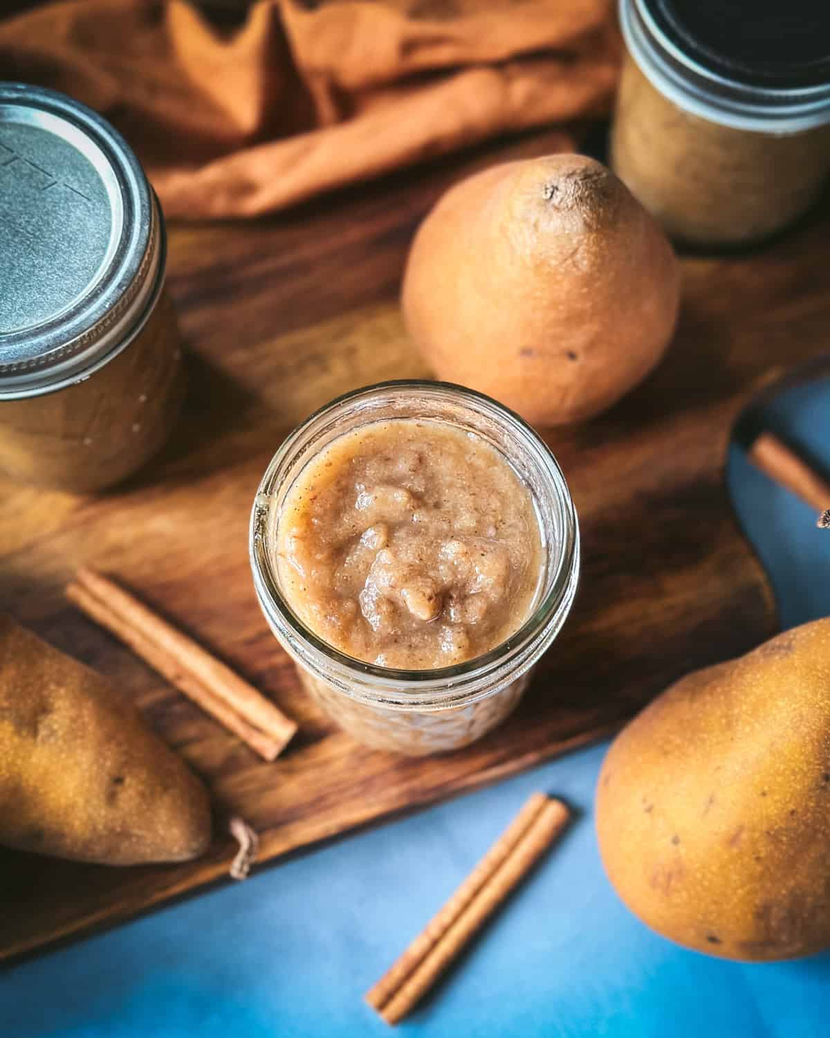 A jar of pear butter surrounded by whole pears and cinnamon sticks, top view. 