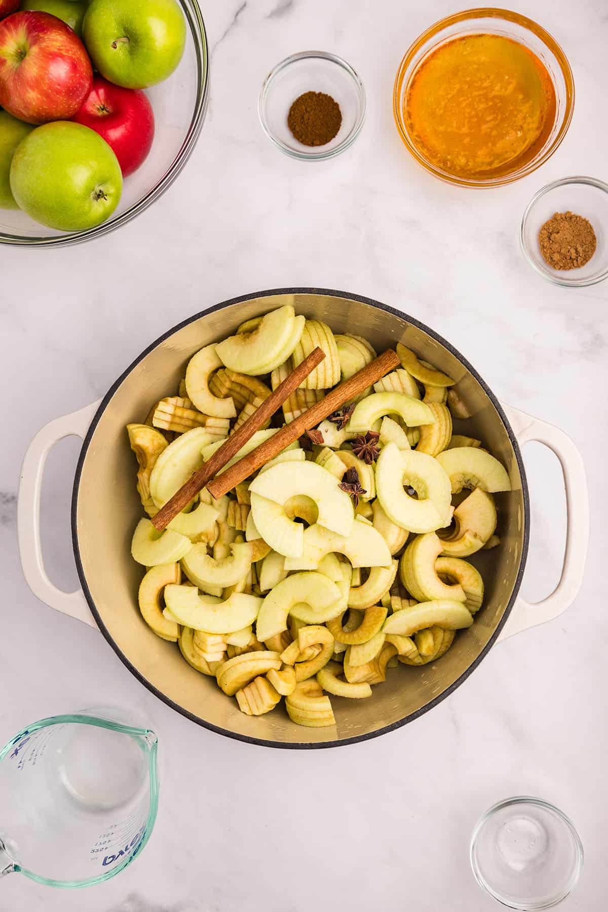 Peeled and cut apples in a pot with cinnamon sticks, with a bowl of fresh apples and smaller bowls of other ingredients surrounding. Top view. 