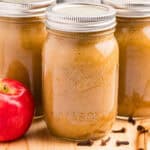 3 jars of canned applesauce on a wood board surrounded by whole cloves and a fresh apple.