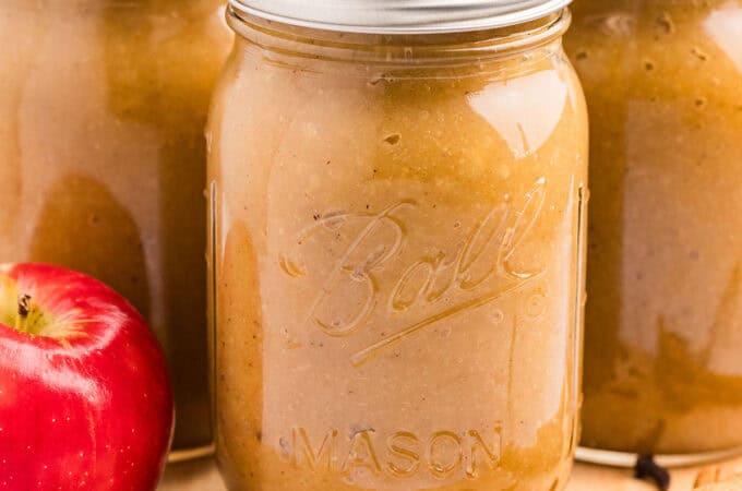 3 jars of canned applesauce on a wood board surrounded by whole cloves and a fresh apple.