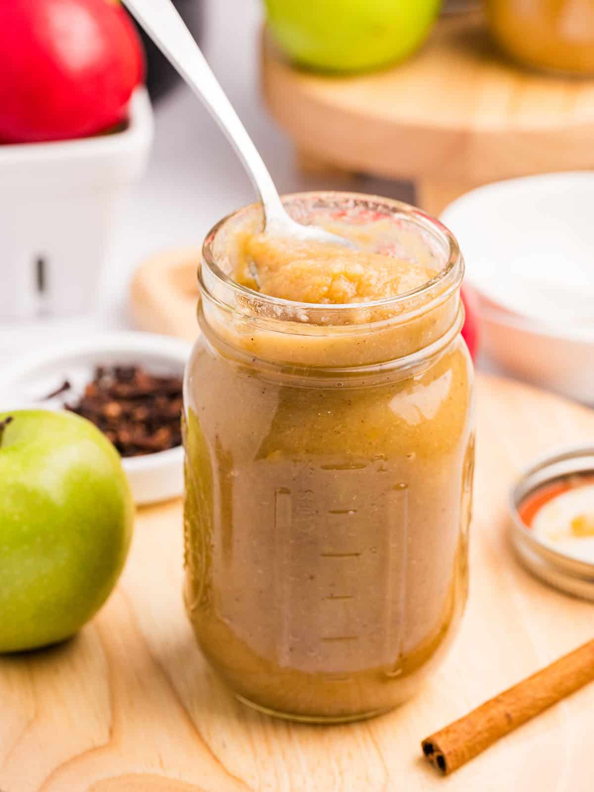 A jar of applesauce with a spoon in it, on a wood surface with other ingredients surrounding. 