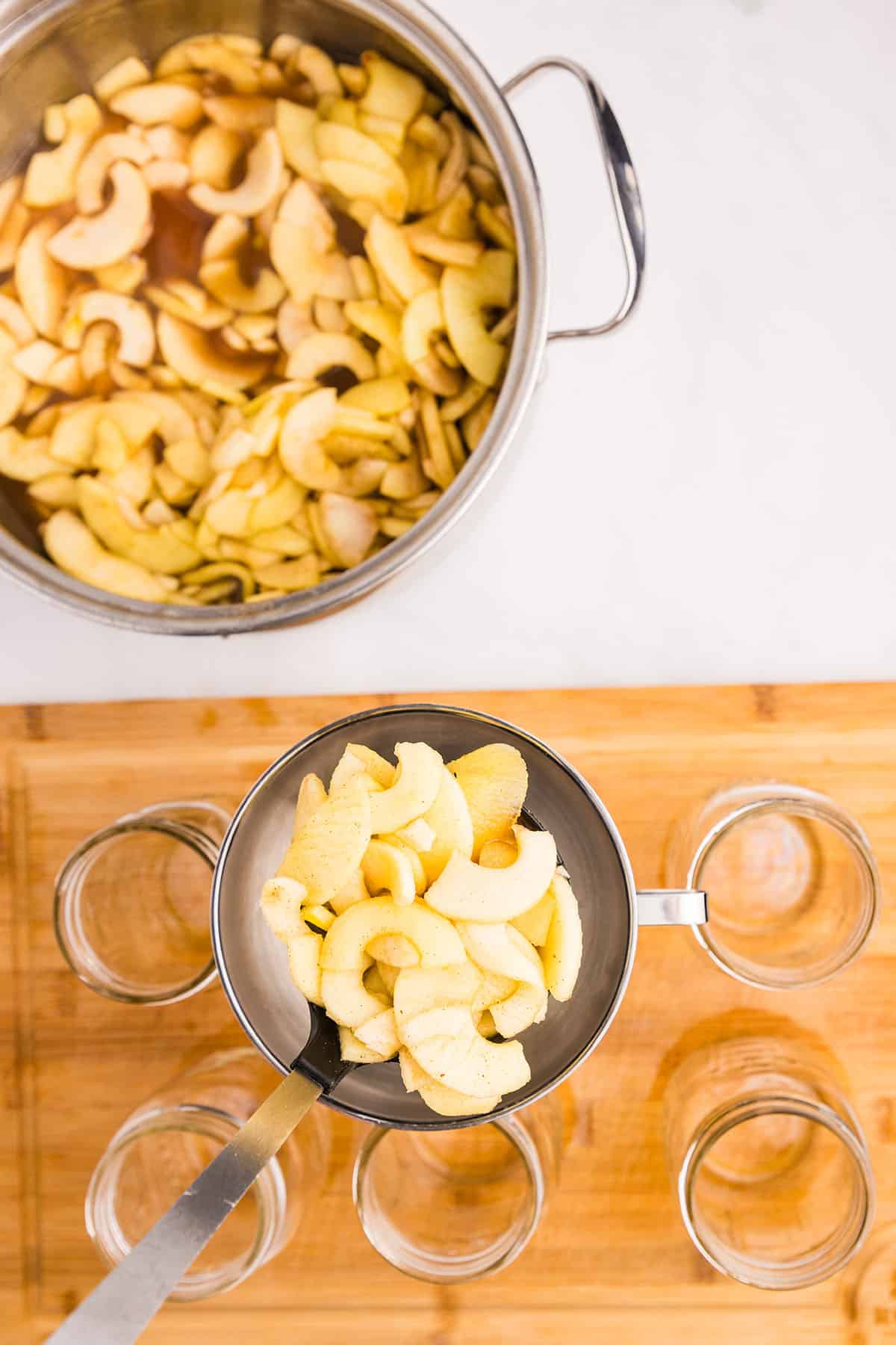 A pot of apple pie filling with a ladle pouring it into prepared jars that are on a wood surface. Top view. 