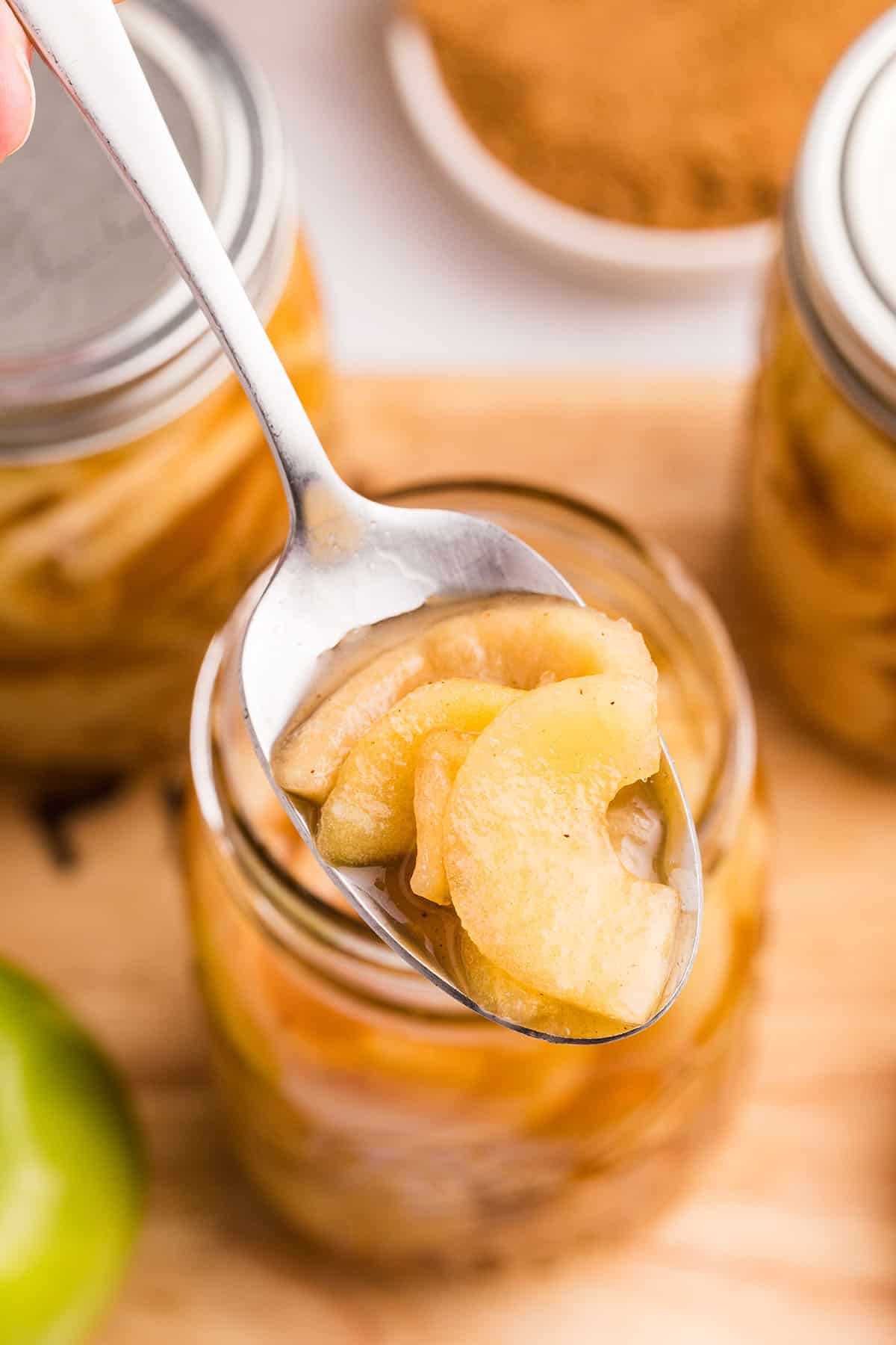 A spoon lifting apple pie filling out of a jar, on a wood surface, top view. 