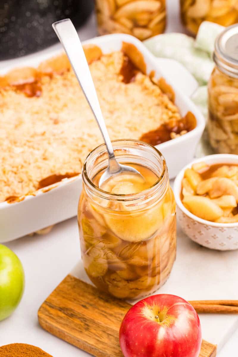 A jar of apple pie filling with a spoon in it, and a crust ready to use for a crumble in the background, surrounded by fresh whole apples.