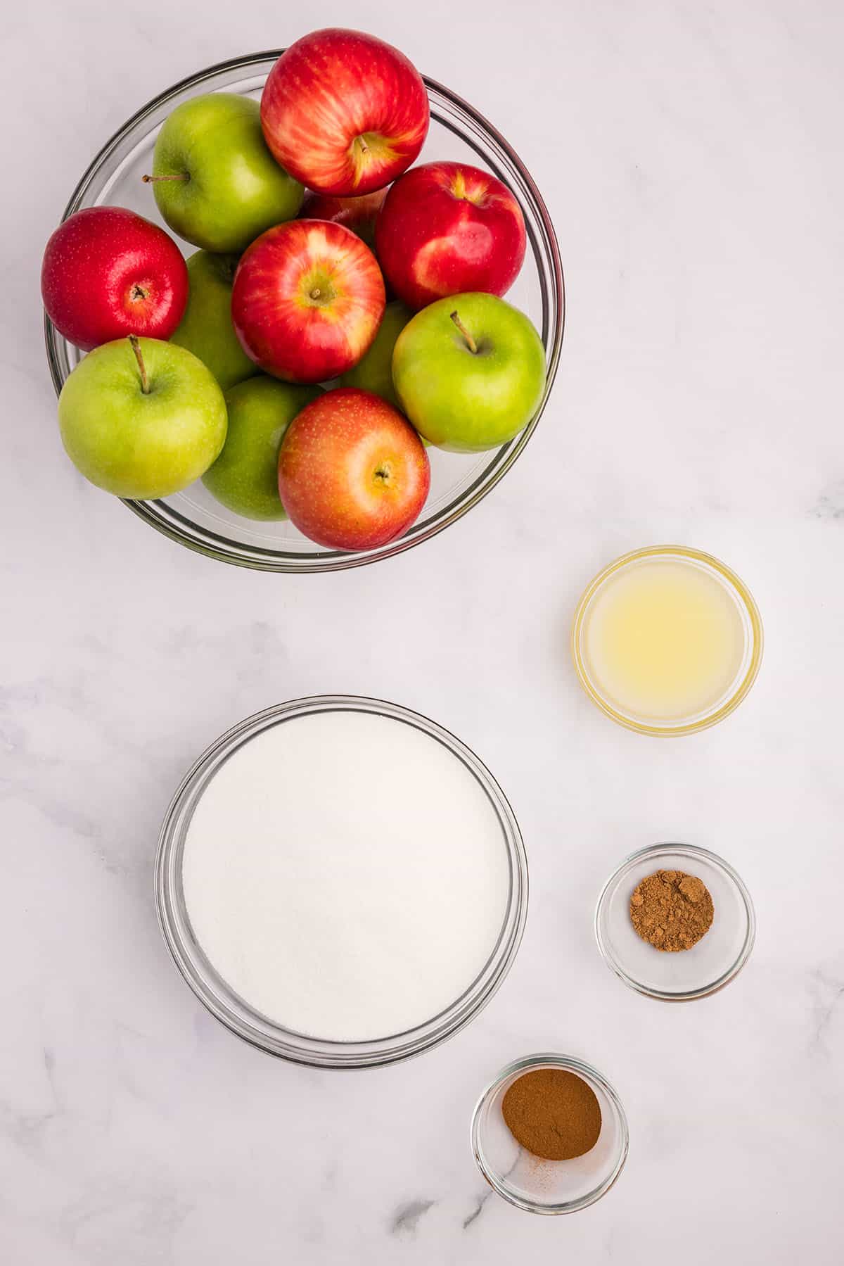 A bowl of mixed apples on a white counter, with smaller bowls of other ingredients, top view. 