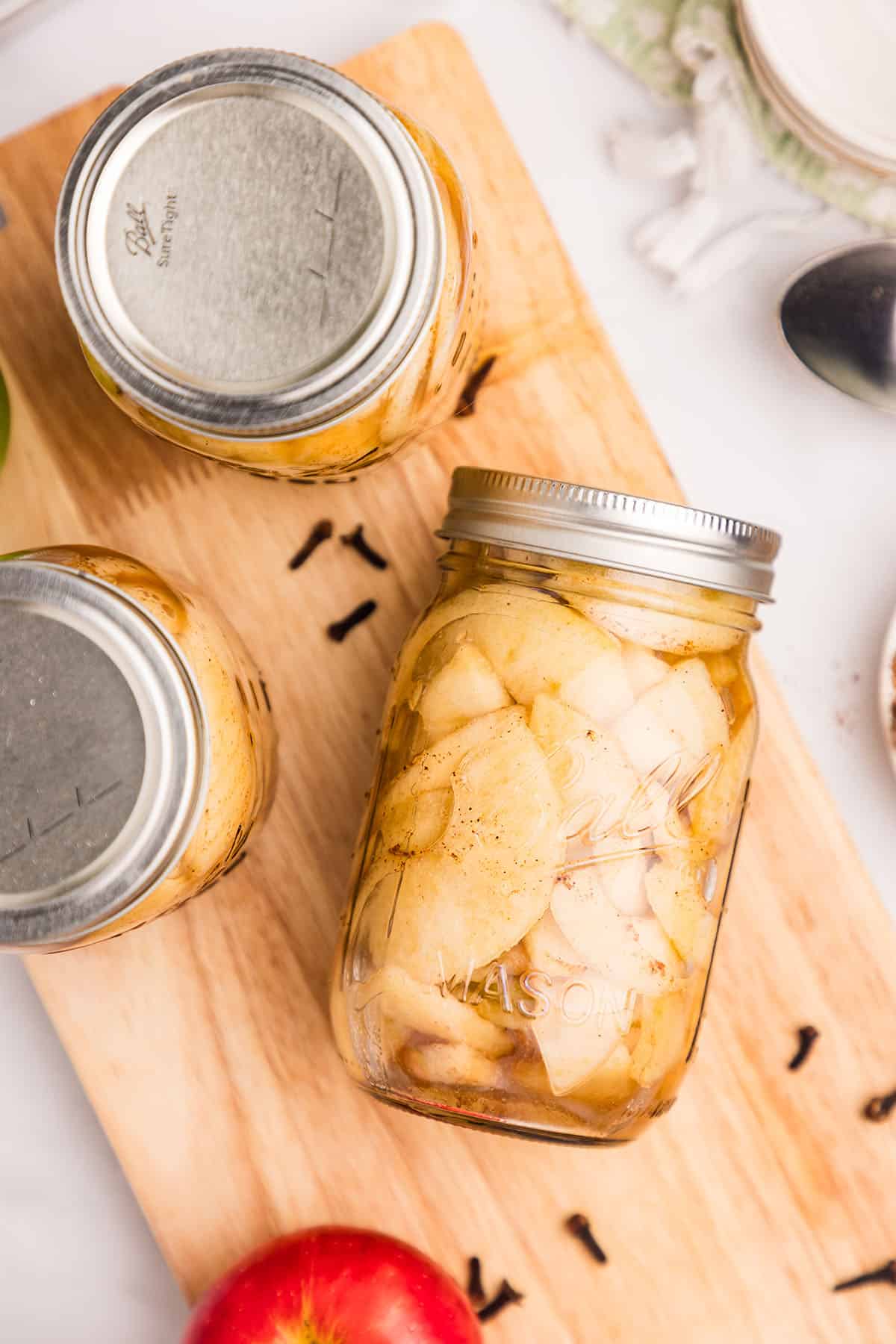 A jar of apple pie filling in it's side on a wood surface, surrounded by whole cloves, apples, and other jars of pie filling. Top view. 