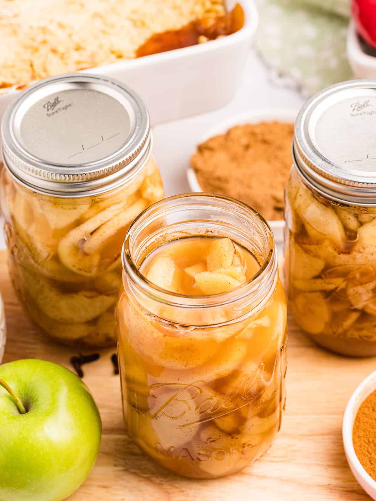 An open jar of pie filling, next to two closed jars. Surrounded by fresh apples. 