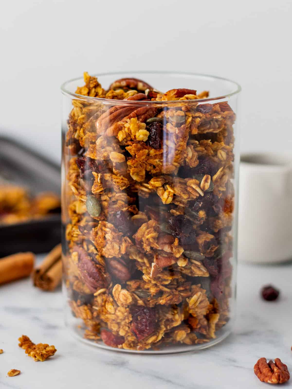 Pumpkin granola in a jar on a white counter with ingredients spread out around it.