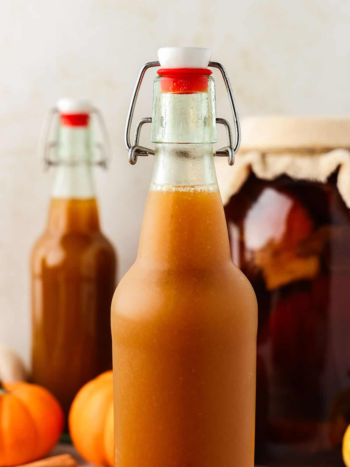 Pumpkin kombucha in a closed bottle, with more in the background and other pumpkins. 