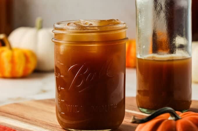 A jar of dark orange pumpkin kombucha with ice, on a wood cutting board surrounded by various pumpkins.