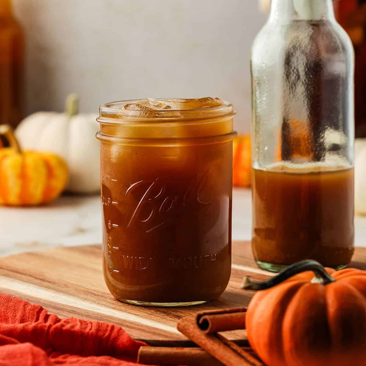 A jar of dark orange pumpkin kombucha with ice, on a wood cutting board surrounded by various pumpkins. 