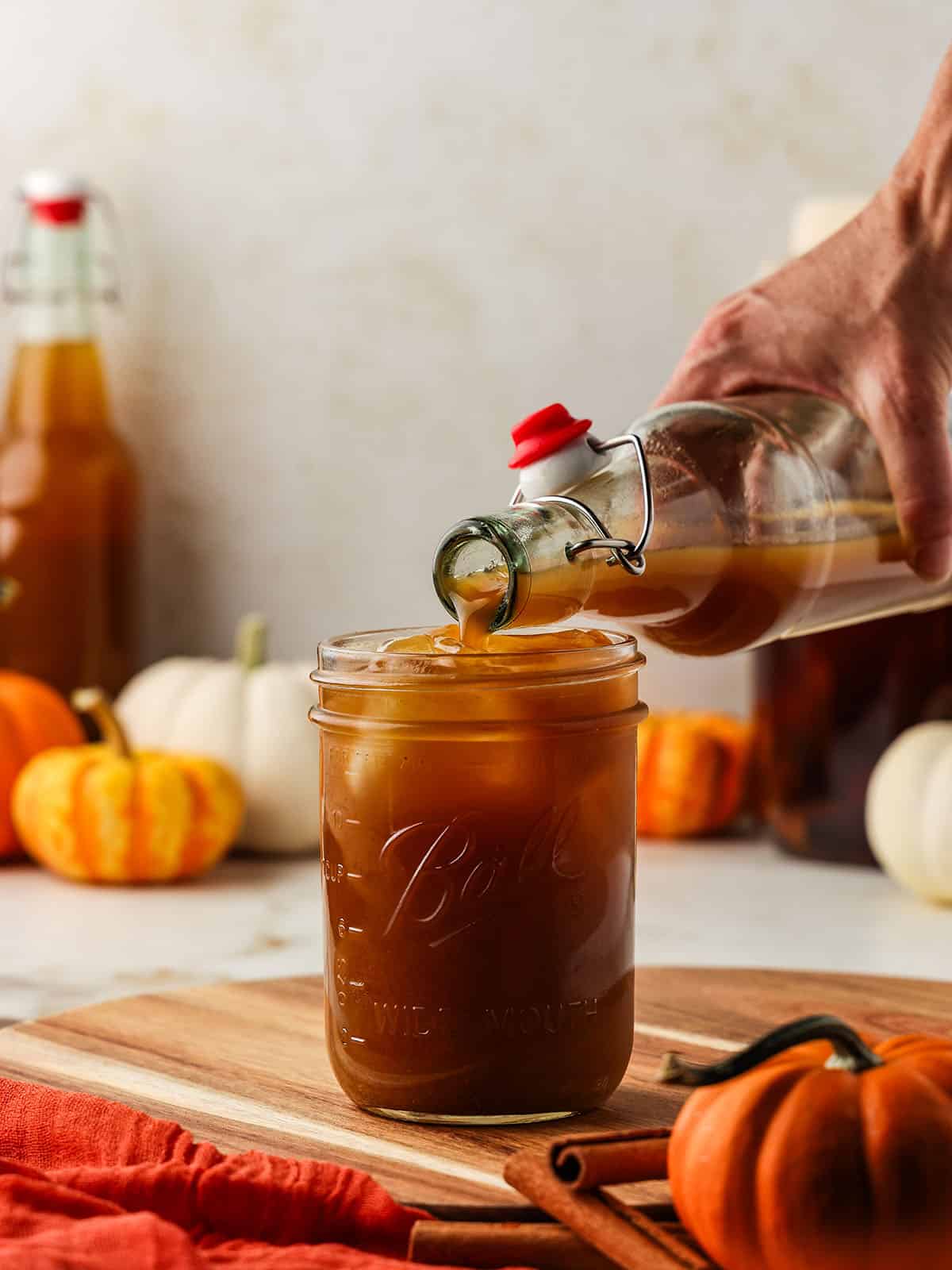 A bottle of pumpkin kombucha pouring into a cup with ice, surrounded by pumpkins. 