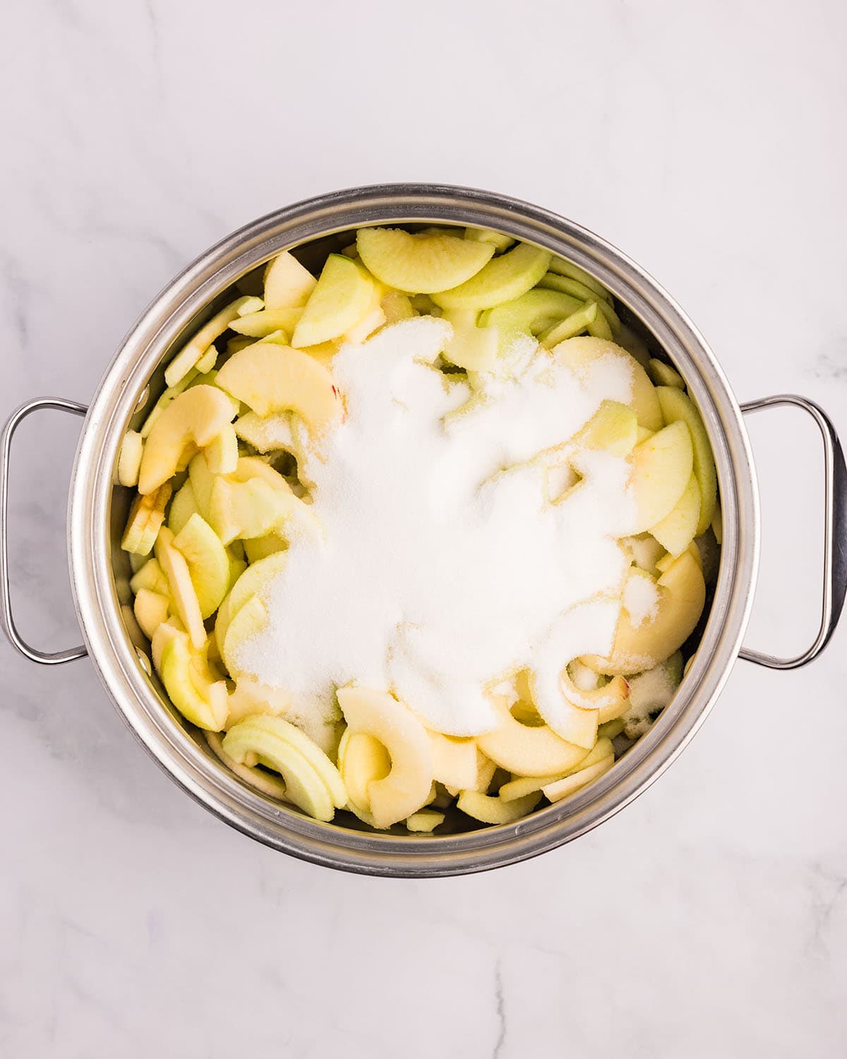 Sliced apples and sugar in a pot on a white surface, top view. 