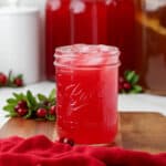 A jar of cranberry kombucha with ice on a wood surface surrounded by a red cloth and fresh cranberry branches.