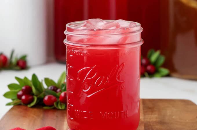 A jar of cranberry kombucha with ice on a wood surface surrounded by a red cloth and fresh cranberry branches.