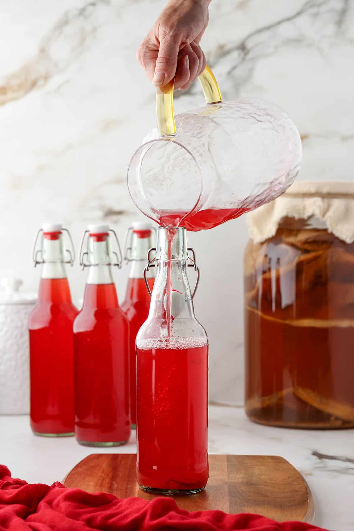 Cranberry kombucha starter pouring from a large pitcher into flip top bottles.