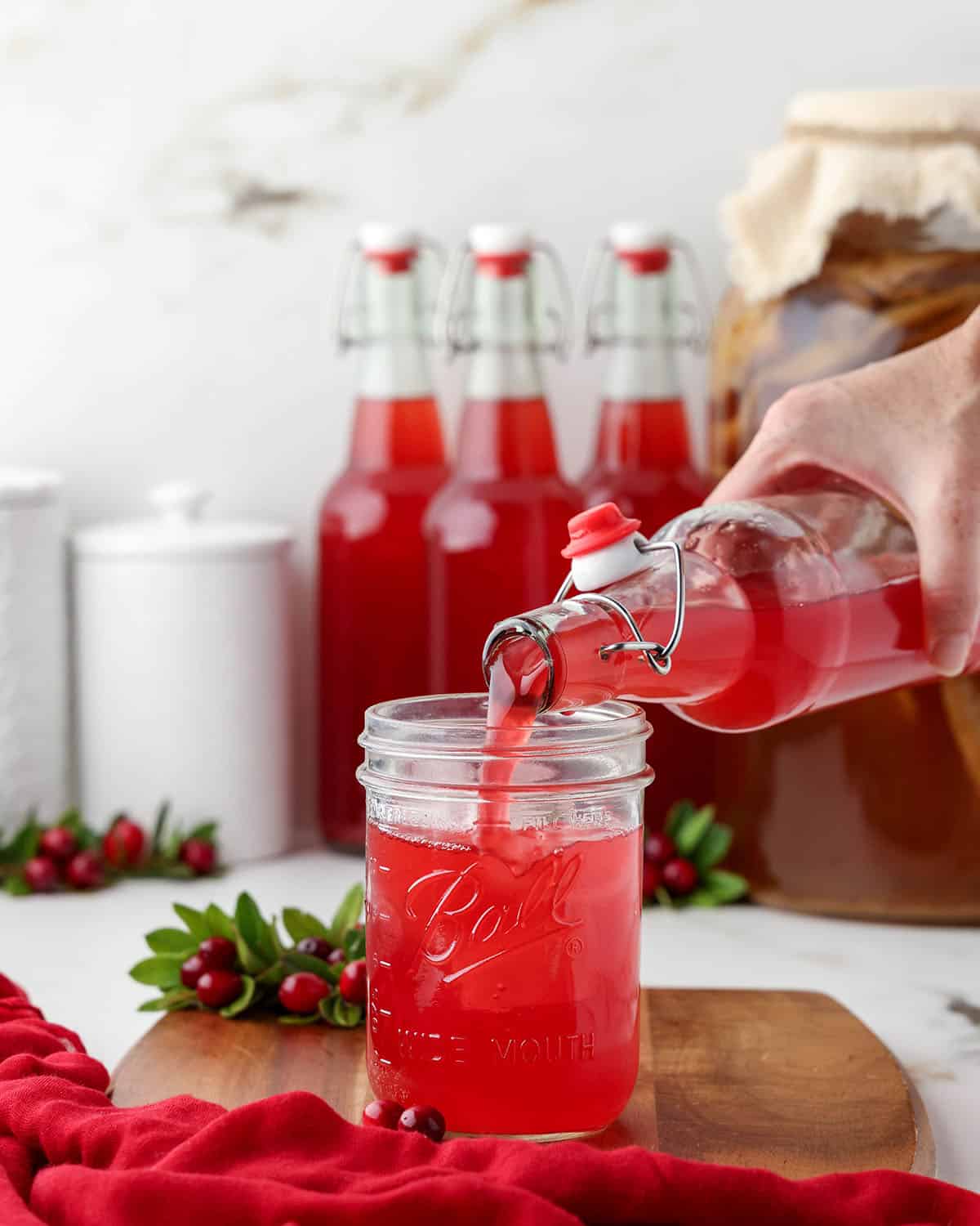 Cranberry kombucha pouring into a glass surrounded by other bottles of kombucha and fresh cranberry branches.