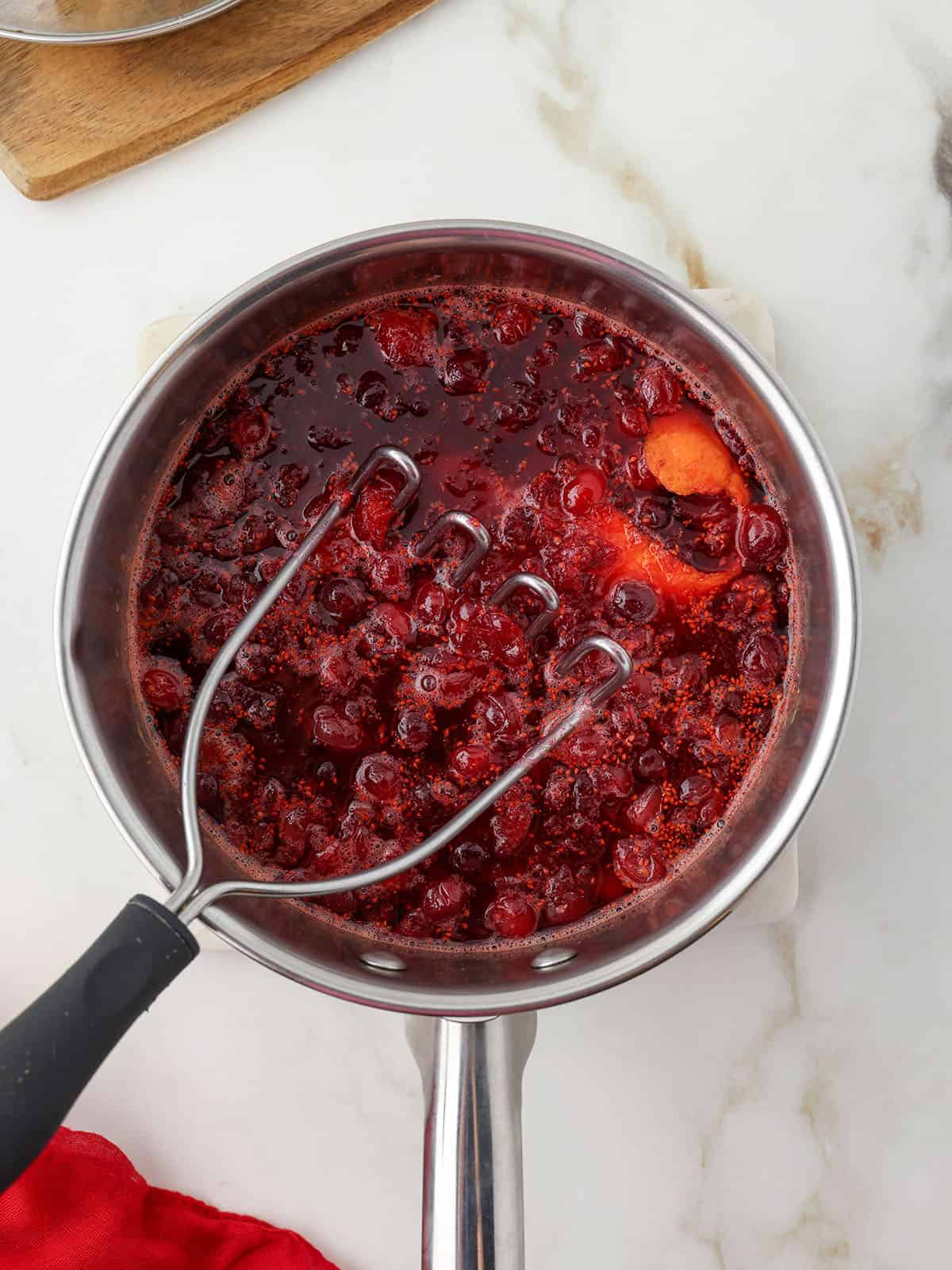 A pot with cranberries being mashed with a potato masher.