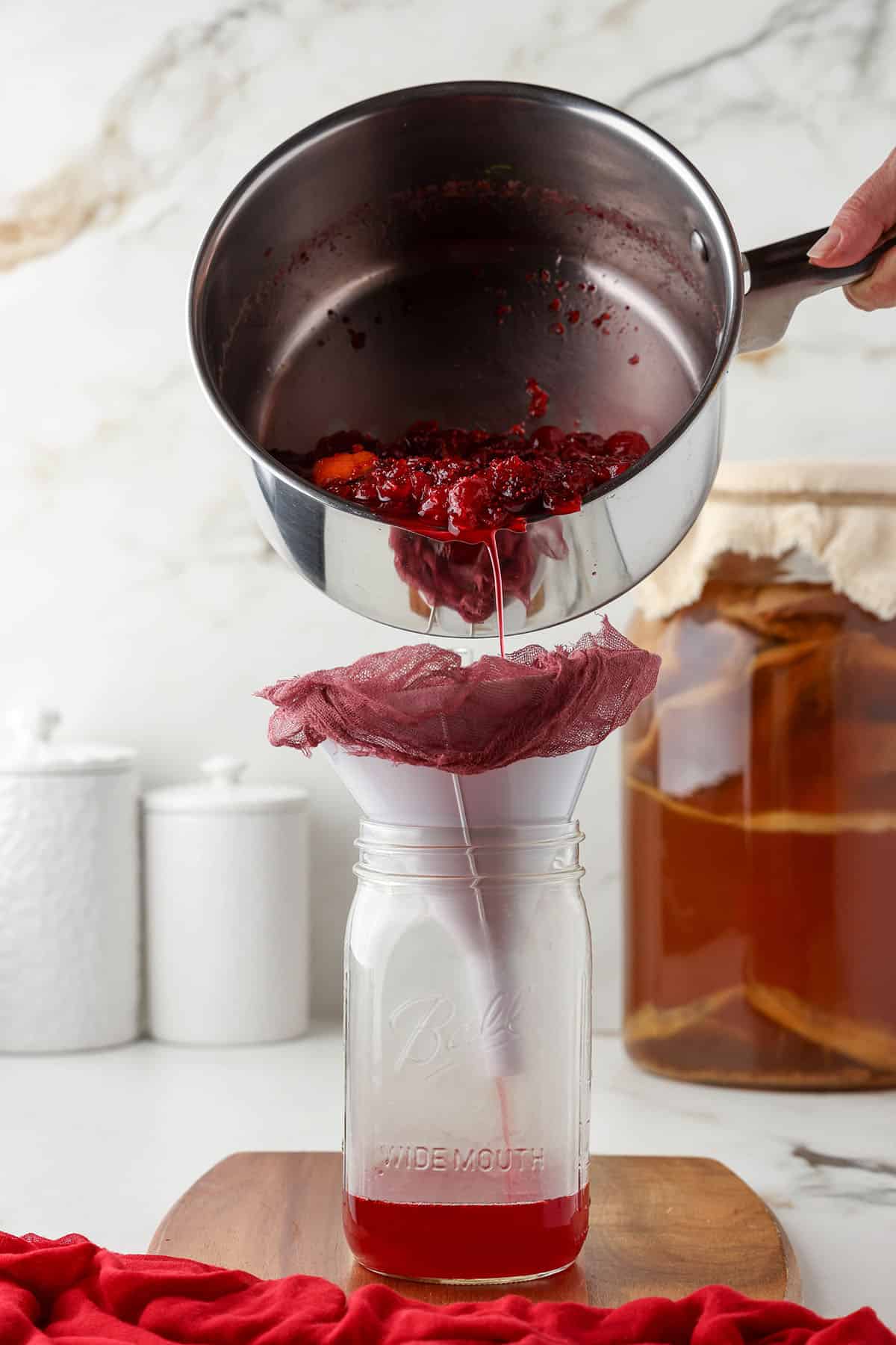 A pot of cranberry syrup pouring through a funnel with cheesecloth into a jar.